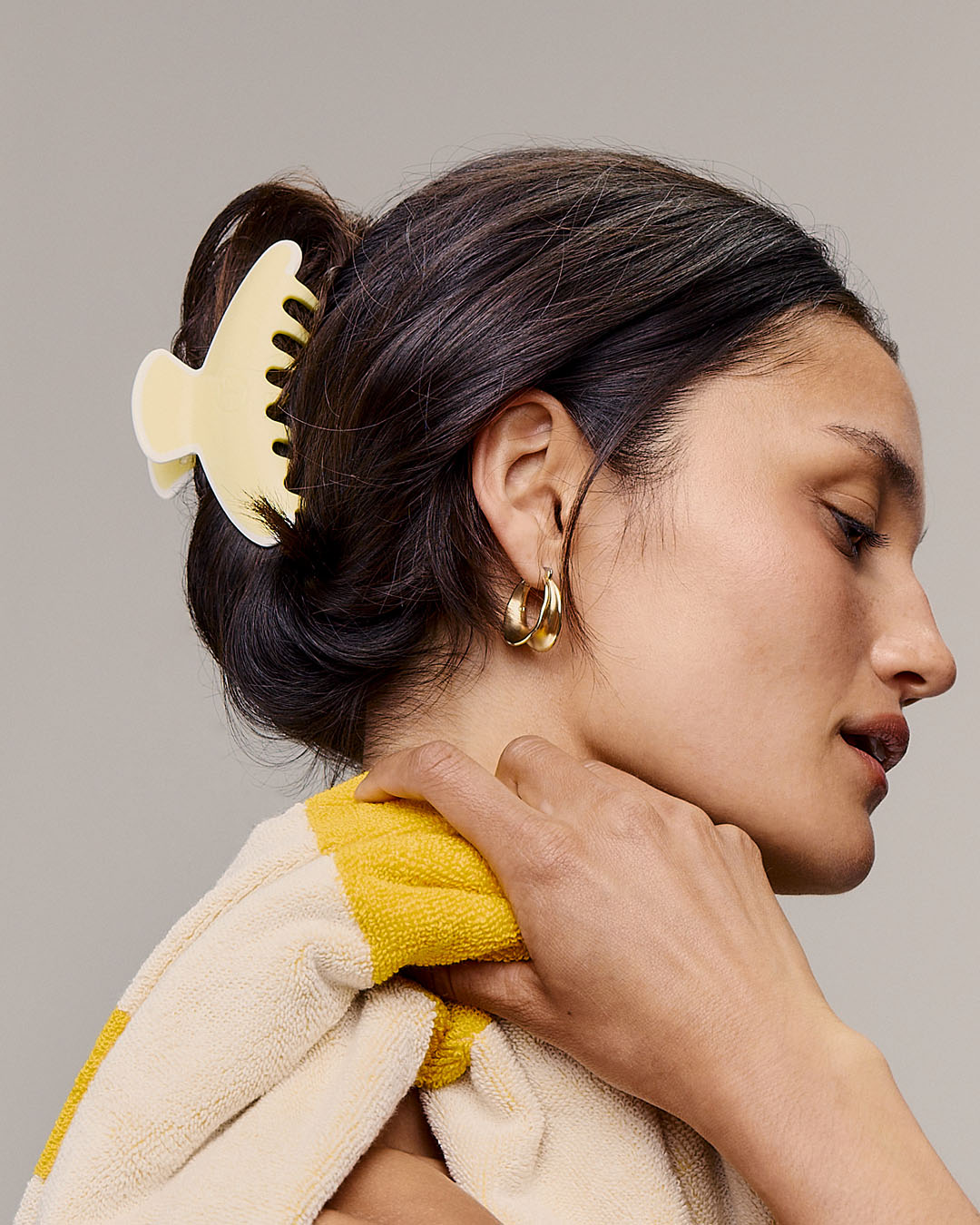Woman with a yellow hair clip and towel, wearing gold earrings, against a neutral background